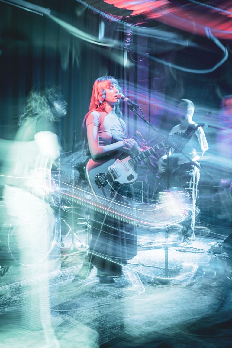Female musician singing and playing guitar live on stage with band, captured with long exposure light trails and vibrant teal stage lighting effects.